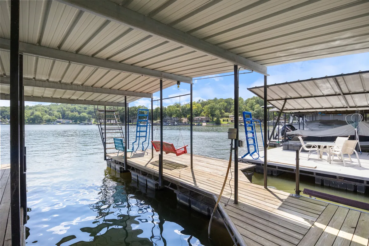 A tranquil wooden dock extends over the calm lake, outfitted with a row of colorful chairs, perfect for relaxing, launching paddle boards, or gathering before a swim. The serene water and tree-lined shore suggest a private, peaceful cove ideal for group water activities.