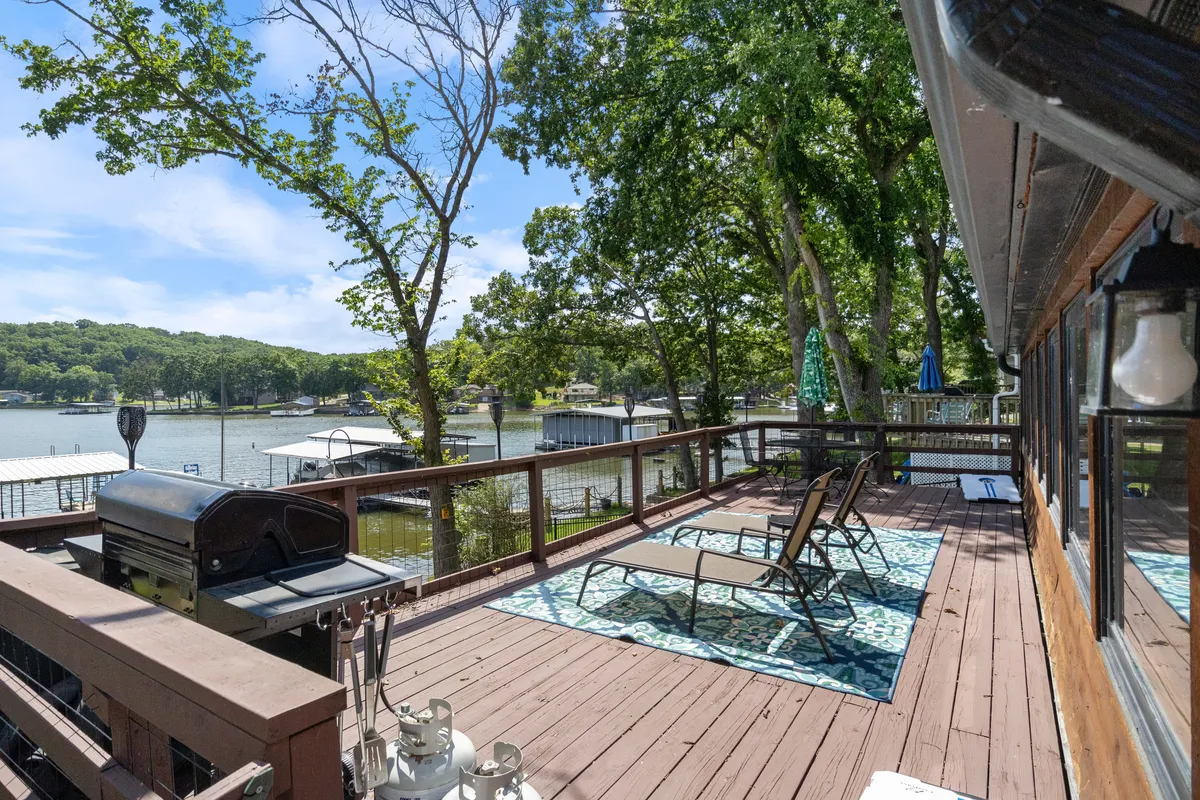 This image presents an inviting exterior deck overlooking the lake, featuring lounge chairs, a grill, and a beautiful natural backdrop with mature trees. The vantage captures the essence of relaxing, dining, and socializing outdoors while immersed in the lakeside atmosphere.