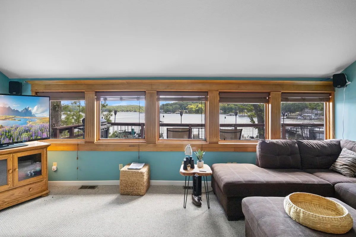 A cozy living room featuring a comfortable sectional sofa positioned in front of large windows that overlook the lake. The space is flooded with natural light, and a TV is prominently displayed for movie nights. Decorative items on the coffee table and a scenic landscape on the TV screen enhance the inviting ambiance.