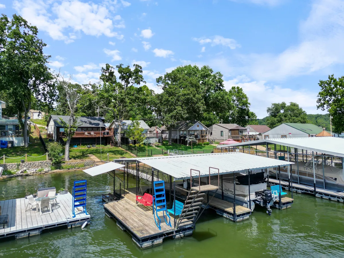 The image showcases a tranquil lakefront setting with a private dock, boathouses, and plenty of outdoor seating, all framed by lush trees. The sun is either rising or setting over the calm water, hinting at beautiful views and peaceful moments. The outdoor areas invite gatherings, fishing, or simply relaxing by the water's edge.