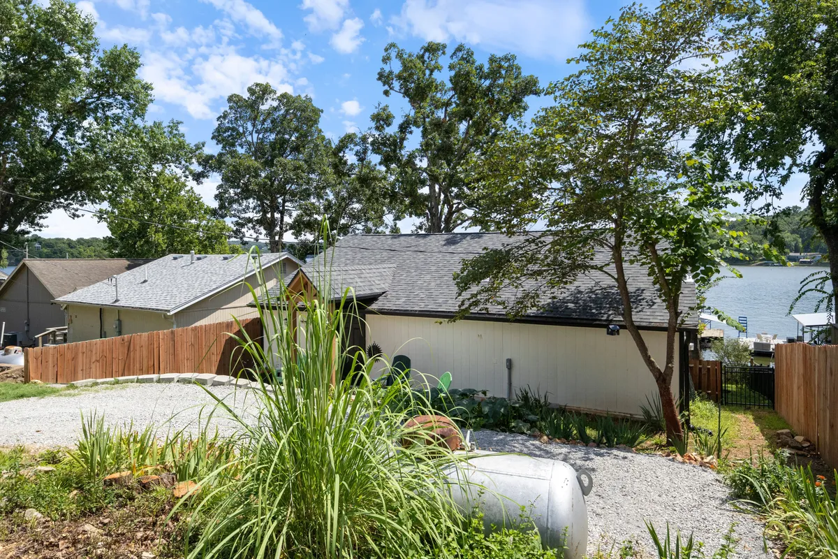 This exterior image features a gravel driveway leading to a fenced backyard with lush greenery and a partial water view. The scene is relaxed and inviting, with open outdoor areas that would appeal to pet owners seeking secure, comfortable spaces for their pets to explore and relax in nature.