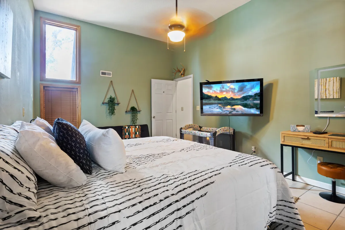 A cozy bedroom featuring a bed with striped bedding, a wall-mounted TV, decorative plants, and wooden furniture. Natural light filters in, creating an inviting and restful environment, ideal for both relaxation and unwinding after a day outdoors.