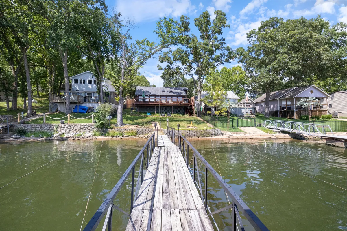 A tranquil scene featuring a wooden dock stretching out over a calm lake, surrounded by lush trees and a few neighboring houses. The dock is the focal point, perfect for launching boats, fishing, or simply relaxing while enjoying the view. The area is quiet and secluded, ideal for outdoor enthusiasts seeking a lakeside retreat.