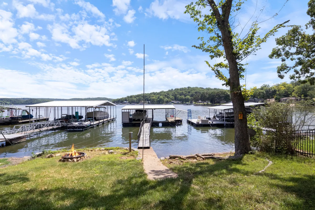 A wooden dock extends into a tranquil waterway, with boats moored nearby, a fire pit area, and trees lining the shore. The scene suggests opportunities for boating, fishing, evening gatherings around the fire pit, and peaceful moments by the water.