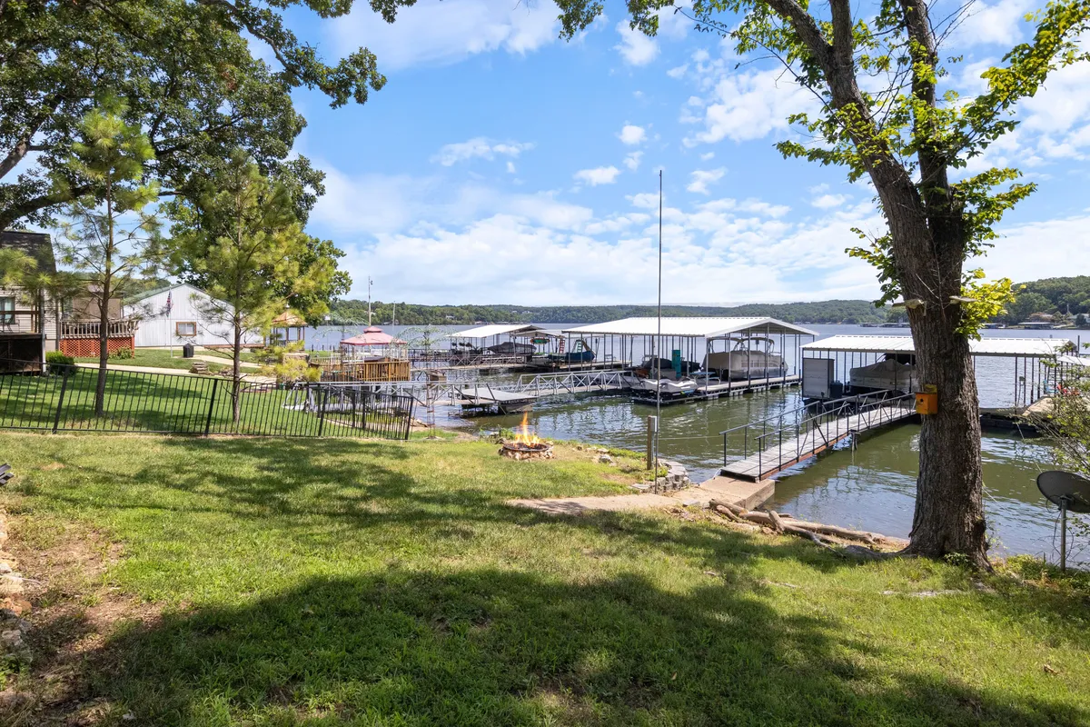 A broad lakeside view showing boathouses, a firepit area, green grass, and trees, all positioned beside the shimmering lake. The scene is calm and inviting, suggesting a setting for boating, fishing, evening bonfires, or simply relaxing by the water at sunset.