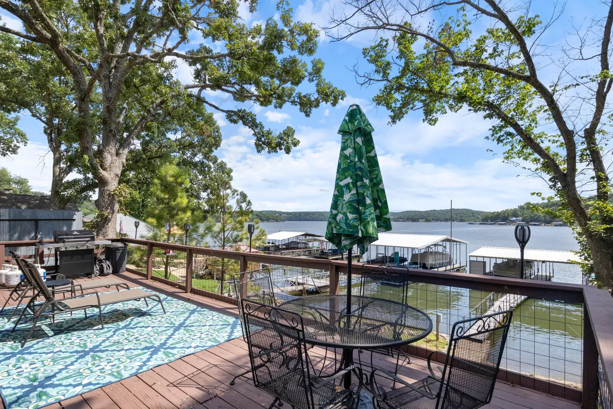 This image showcases an outdoor dining area with a table and chairs under a green umbrella, a decorative rug, and a beautiful view of the lake directly beyond the deck. The space is set up for relaxed al fresco meals, with easy access to the water and dock for swimming or water sports.